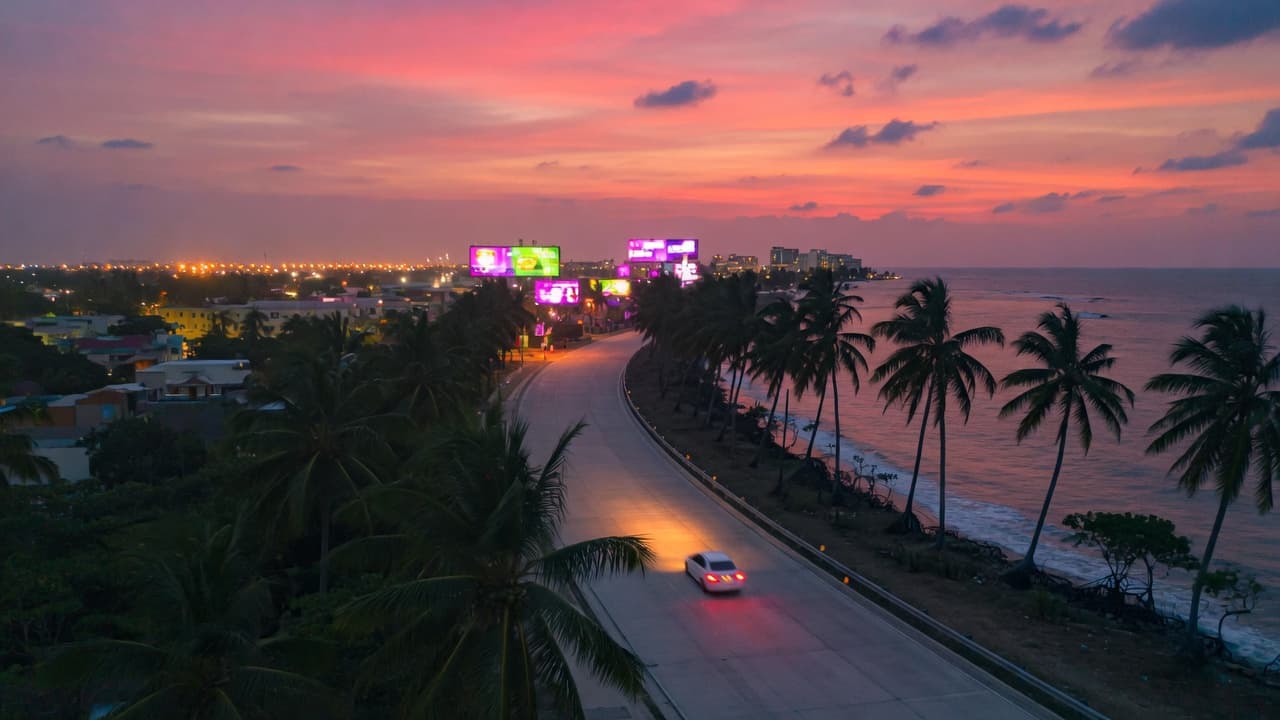 Dusk over a tropical coastal highway heading toward a neon city glow, original BroadbandPicker artwork.