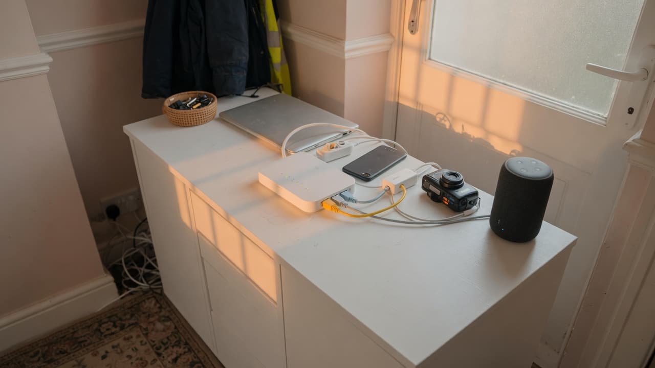 A UK hallway cabinet with a home router, laptop, phone, camera and smart speaker sharing one line, original BroadbandPicker photograph.